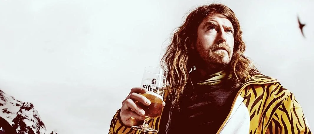 Craig Campbell on a mountain top in a tiger-stripe jacket, raising a beer